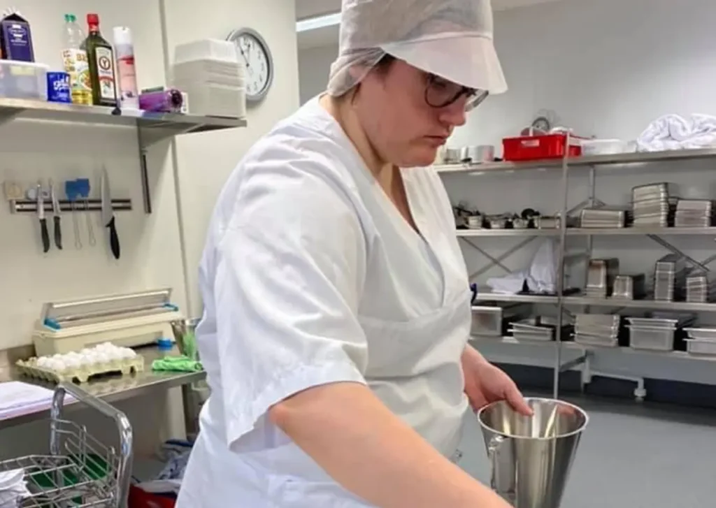 En person på et storkjøkken iført en hvit uniform, caps og briller, holder en bolle i rustfritt stål. Ulike kjøkkenredskaper, ingredienser og redskaper er synlige på hyller og benkeplater mens de ekspert demonstrerer sine kompetansebrev-ferdigheter.