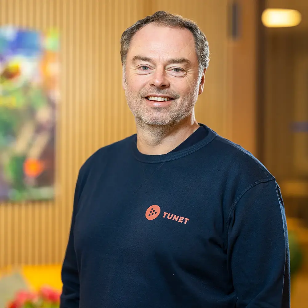 En mann i en marineblå genser med ordet «Tunet» trykket på står innendørs og smiler til kameraet.