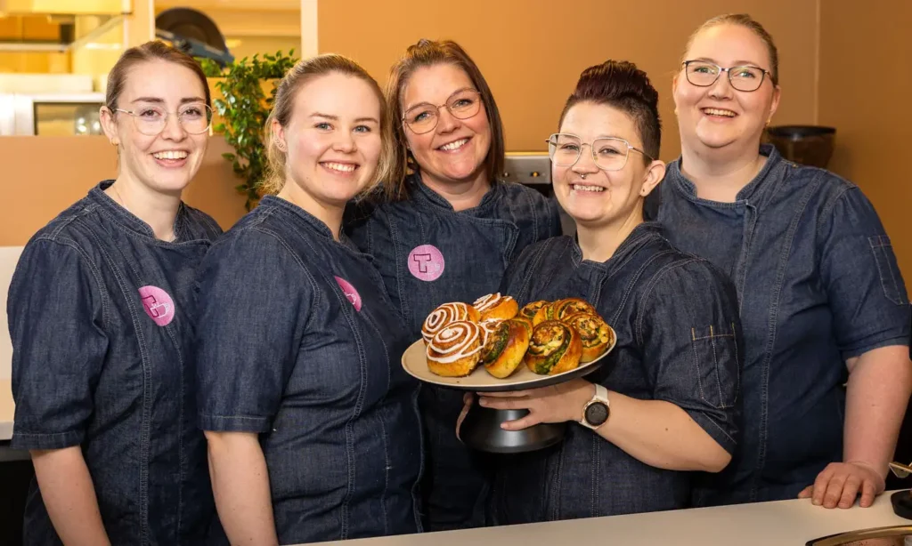 Fem smilende individer i matchende dongeriuniformer står sammen innendørs på Tunet Kafe, en holder et fat med ulike bakevarer.