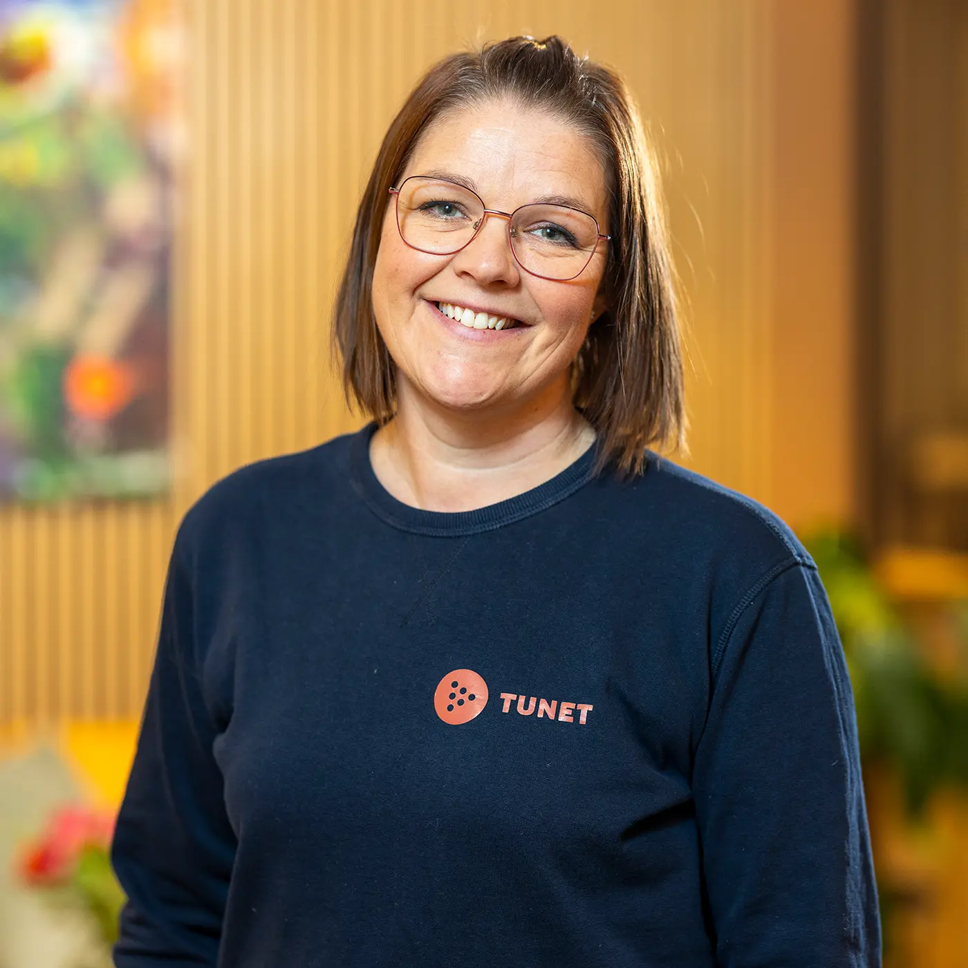 En kvinne med briller og skulderlangt hår smiler. Hun har på seg en marineblå skjorte med logoen «TUNET» trykt på. Bakgrunnen ser ut til å være en innendørs setting med varm belysning, og skaper en innbydende atmosfære som virker perfekt for et "kontakt oss"-øyeblikk.