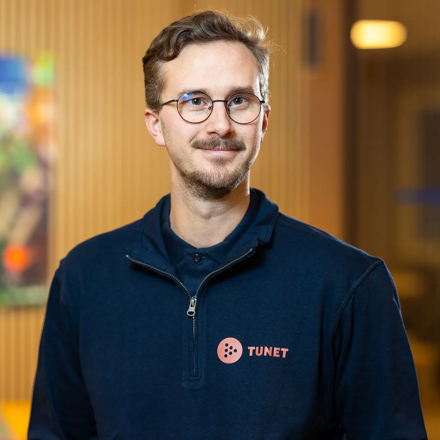 En mann med briller og bart har på seg en marineblå glidelåsjakke med «TUNET»-logo. Han står innendørs med uskarp bakgrunn og virker nærgående. Ta gjerne kontakt eller kontakt oss for eventuelle spørsmål.