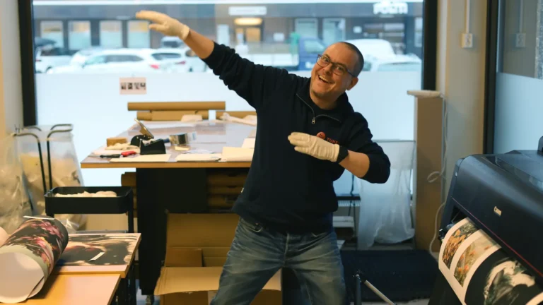 En mann med hansker poserer lekent med en "dab"-gest på et kontor eller trykkeri, omgitt av skrivebord og trykksaker - et øyeblikk fra "Fra avgrunn til fagbrev - Kims inspirerende reise tilbake til livet.