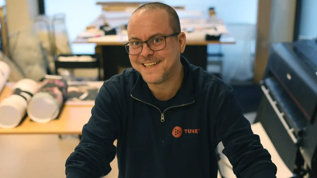 En mann med briller og marineblå «TUNE»-genser smiler mens han sitter på et kontor og reflekterer over «Fra avgrunn til fagbrev - Kims inspirerende reise tilbake til livet» med papirer og utstyr i bakgrunnen.