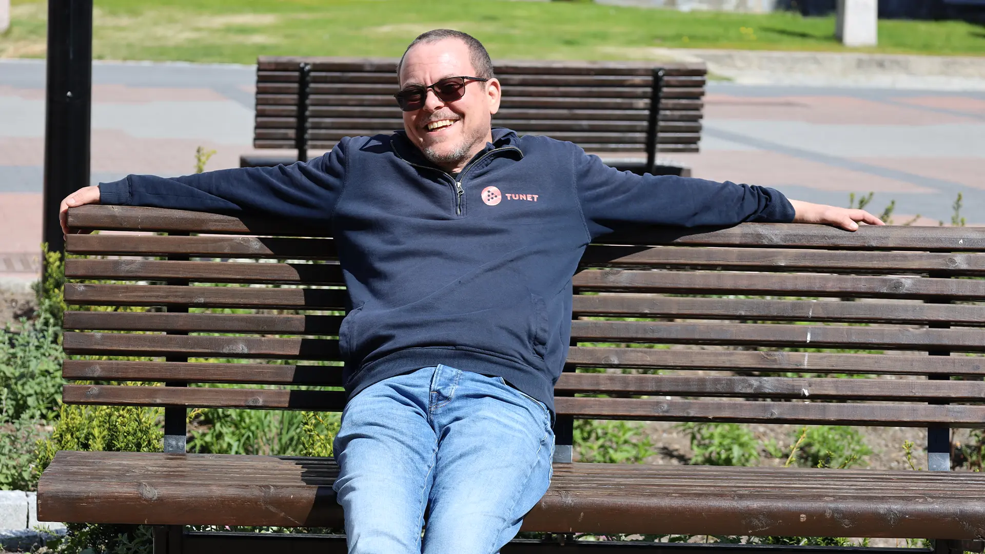En mann med solbriller og marinegenser sitter avslappet på en parkbenk i tre med armene utstrakt og smiler i sollyset – og reflekterer Fra avgrunn til fagbrev - Kims inspirerende reise tilbake til livet.