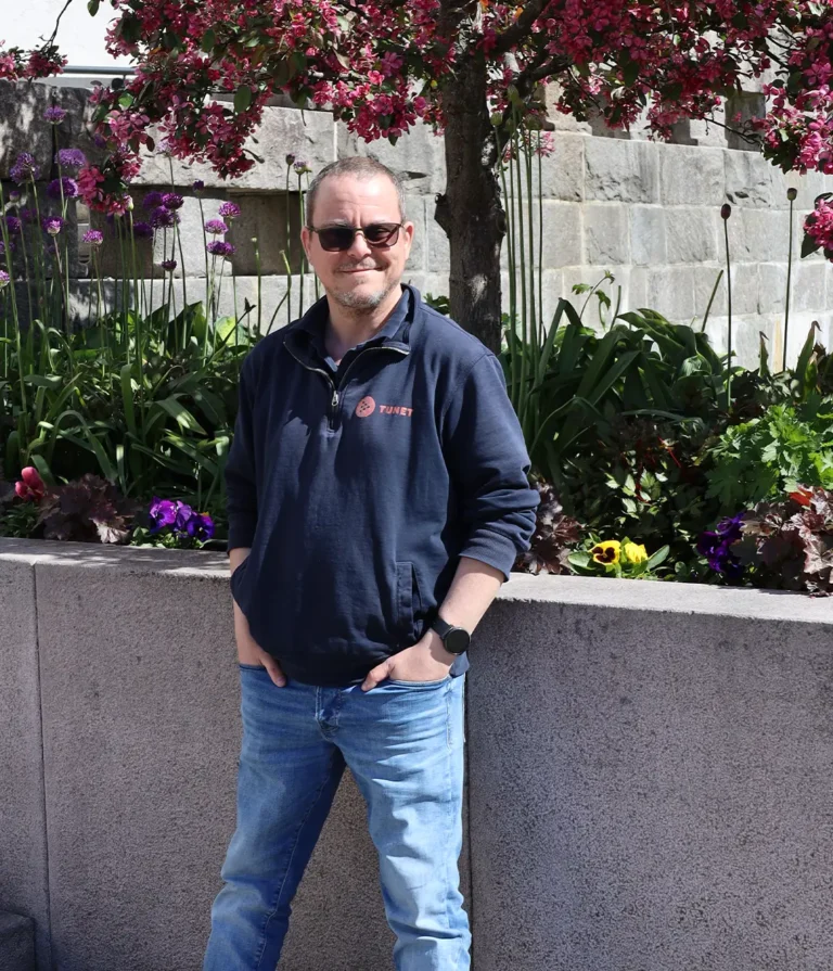 En mann med solbriller, en mørkeblå genser og jeans står utendørs foran en betongvegg og blomstrende planter ved fagbrevtunet.