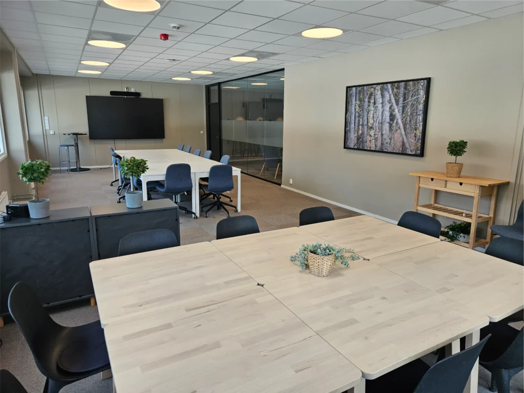 Et moderne konferanserom med lyse trebord, svarte stoler, flere potteplanter, en stor skjerm og et innrammet bilde av trær på veggen – perfekt for møter med Årets samarbeidspartner 2024: Fysen kafé på Terningen Arena.