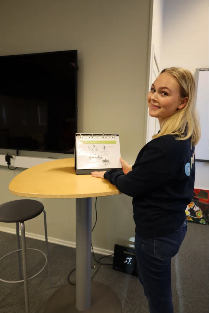 En kvinne står ved et rundt bord, holder en flippover med illustrasjoner, ved siden av en krakk og en stor monitor på veggen.