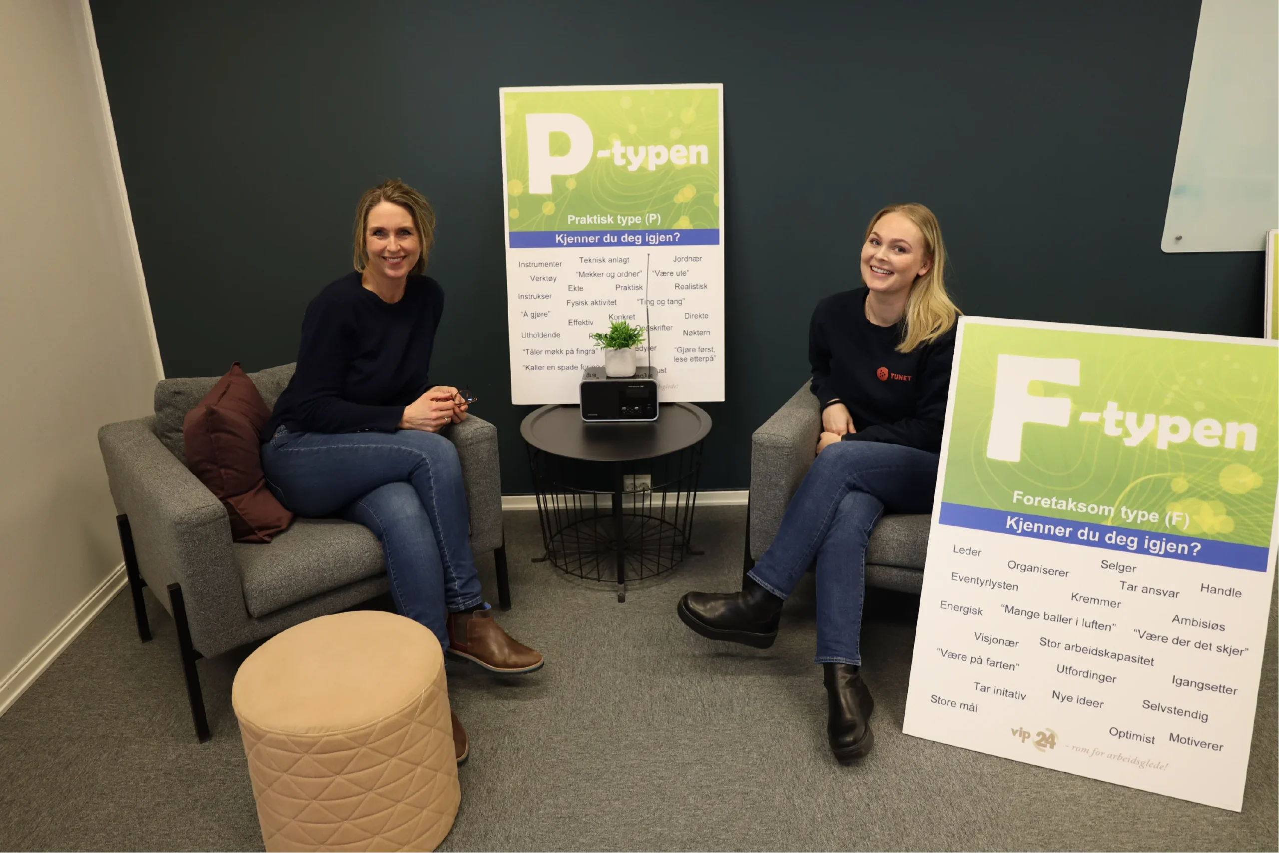 To kvinner sitter i stoler på et kontor og smiler til kameraet. Mellom dem er det et lite bord, og bak dem er det plakater merket «P-typen» og «F-typen».