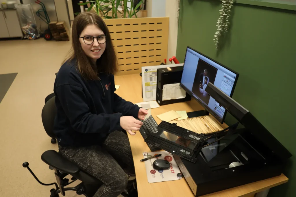 En person sitter ved et skrivebord ved hjelp av en planskanner med ulike papirer og en datamaskin som viser et skannet bilde—gjenoppdag minne dine fra fotograf Westjordet.
