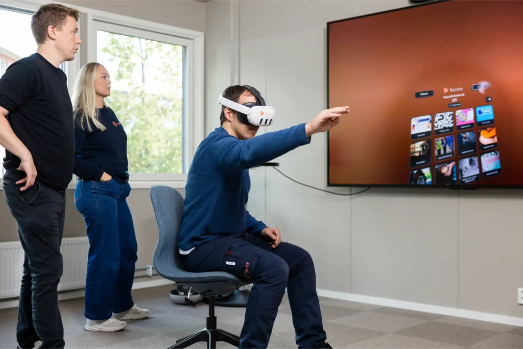 En sittende person med VR-hodesett peker mot en stor skjerm som viser digitalt innhold, mens to andre står i nærheten og ser på.