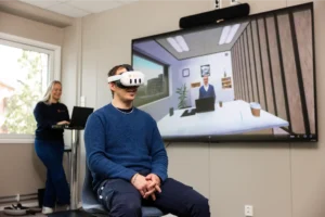 En person med VR-hodesett sitter foran en skjerm som viser et virtuelt kontormøte, helt oppslukt i VR-miljøet, mens en annen person betjener en bærbar PC i bakgrunnen.