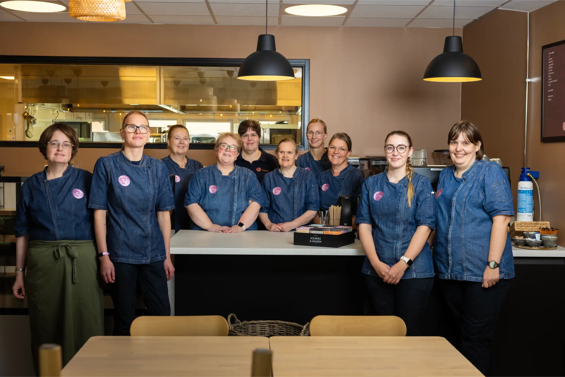 Ti personer i matchende blå uniformer står sammen bak en disk i den moderne Tunet Kafe og poserer for et gruppebilde i det stilfulle kjøkkenmiljøet.