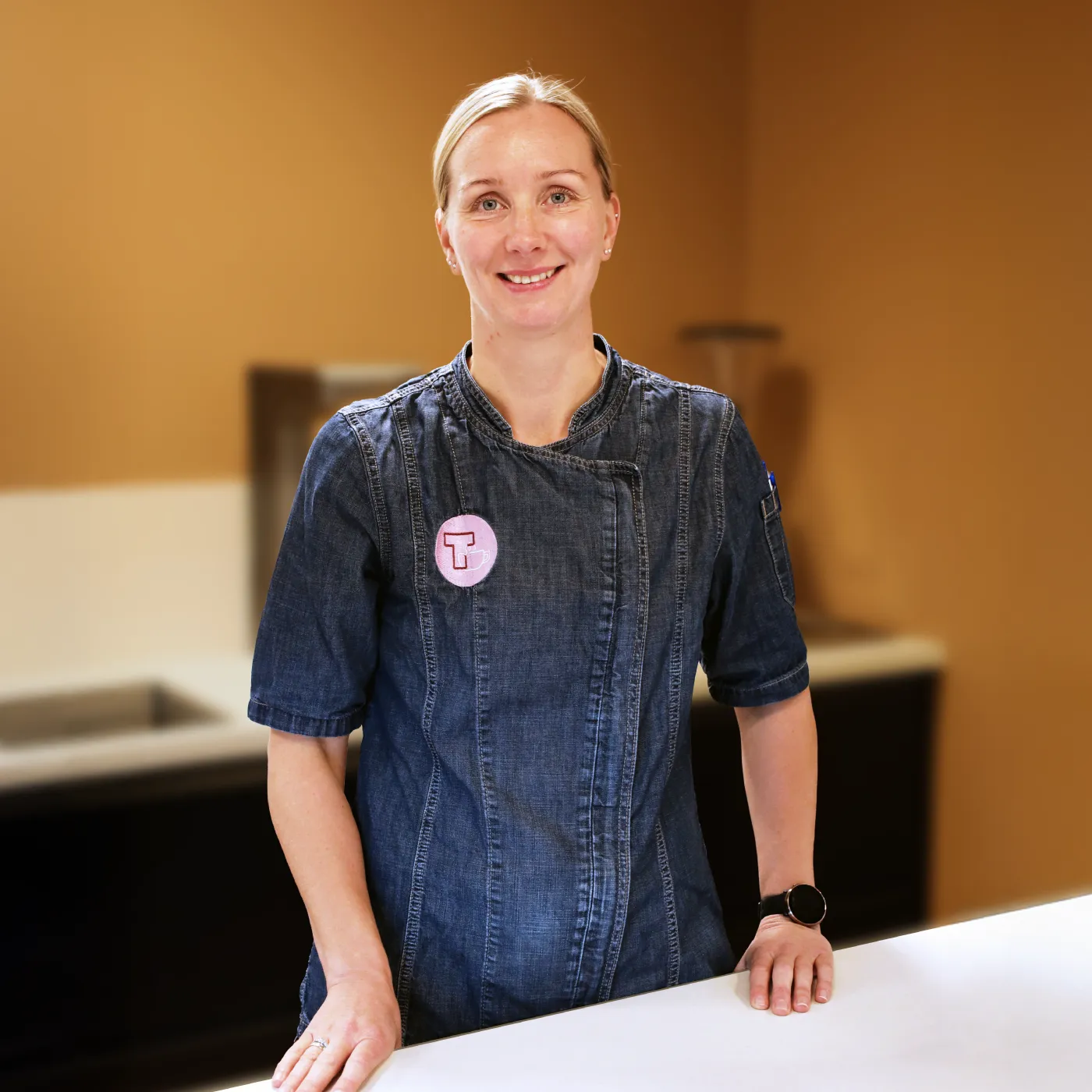 En person iført kokkefrakk i denim og en rund nål står bak en hvit kjøkkenbenk og smiler til kameraet. For henvendelser eller for å lære mer, kontakt oss.