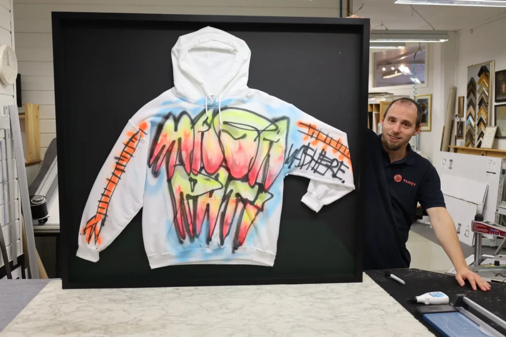 En mann står ved siden av en innrammet hvit hettegenser med objektinnramming i form av fargerik spraymaling i graffitistil og dristige bokstaver i svart, oransje og grønt.