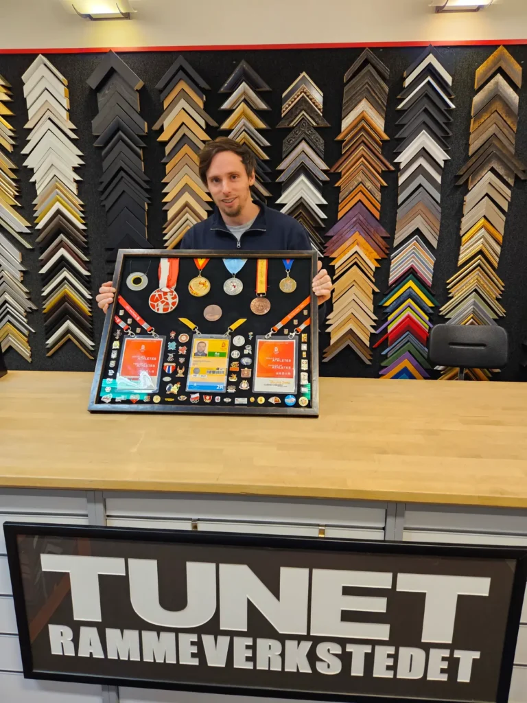 En person står bak en disk med medaljer og memorabilia i en innrammingsbutikk som spesialiserer seg på objektinnramming, med rammeprøver på veggen.