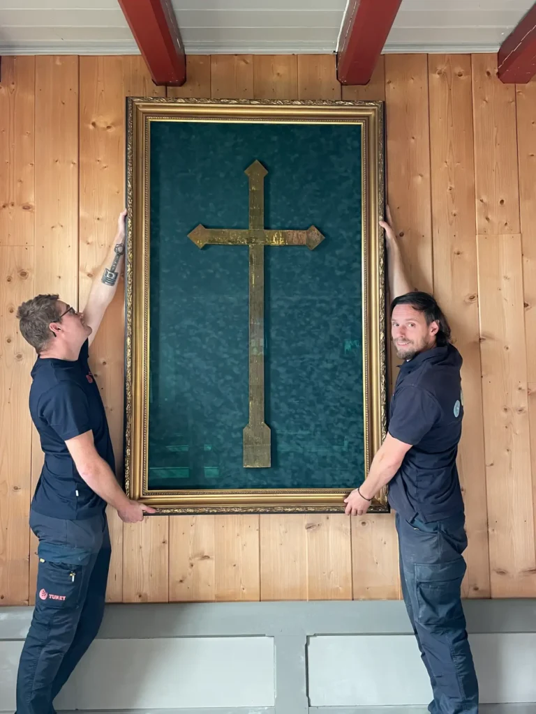 To personer i arbeidsuniformer holder opp et stort, innrammet kunstverk med et gullkors mot en blågrønn bakgrunn, fagmessig fremstilt med objektinnramming, plassert mot en trevegg.