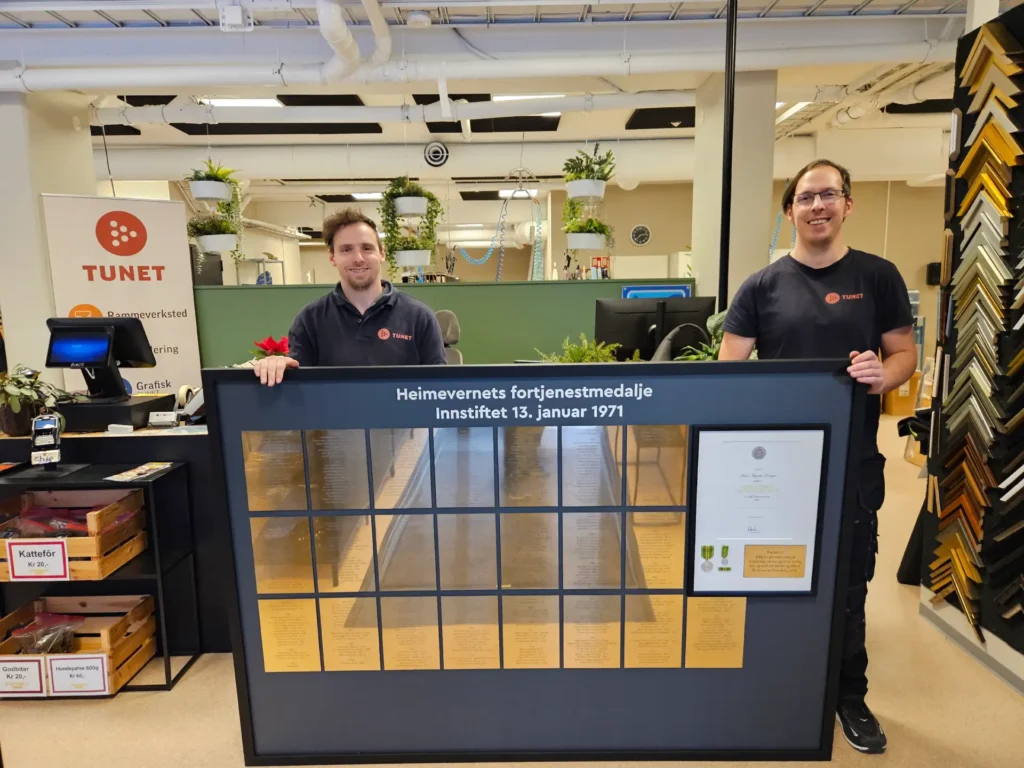 To menn står innendørs, smilende og holder en stor objektinnrammingsmontre med gullplaketter og et dokument. I bakgrunnen skimtes et skilt med teksten "Tunet".