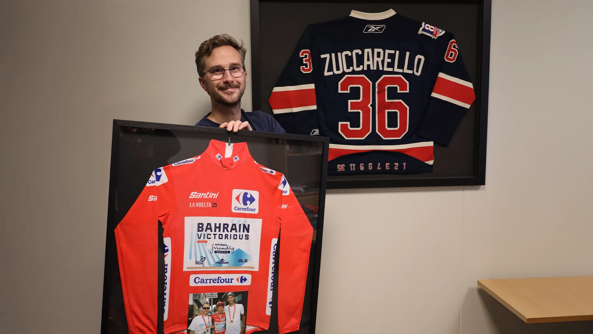 En mann viser frem en innrammet rød sykkeltrøye, med en mesterlig objektinnramming, foran en vegg med en innrammet Zuccarello 36-hockeytrøye.
