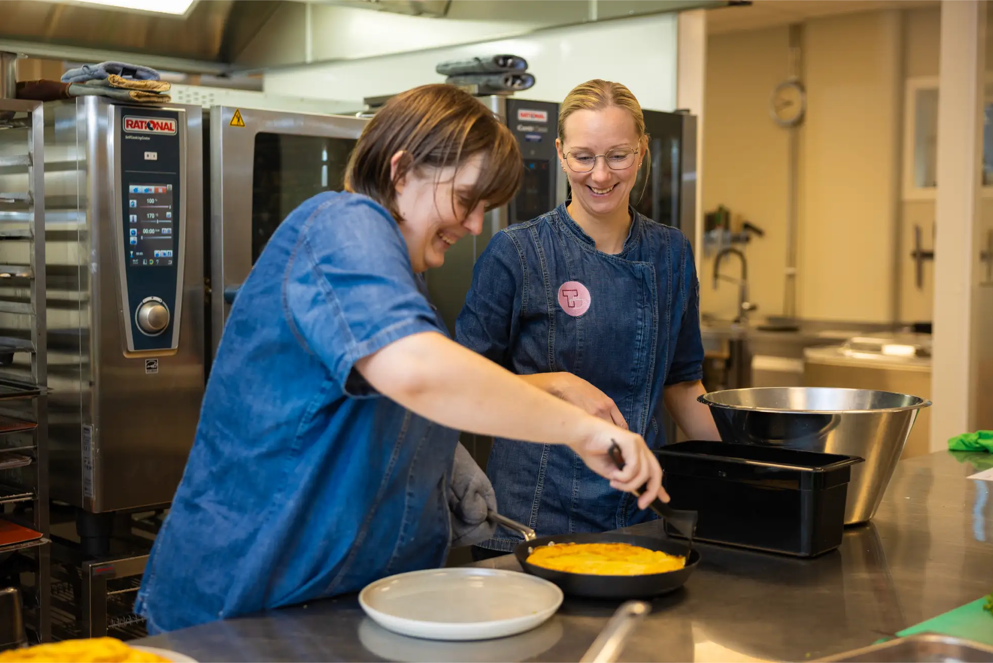 To personer i blå uniformer tilbereder mat sammen på et storkjøkken, mens den ene smilende anretter en rett i en rund panne mens den andre observerer - en innbydende scene for alle som søker en ledig stilling kokk.