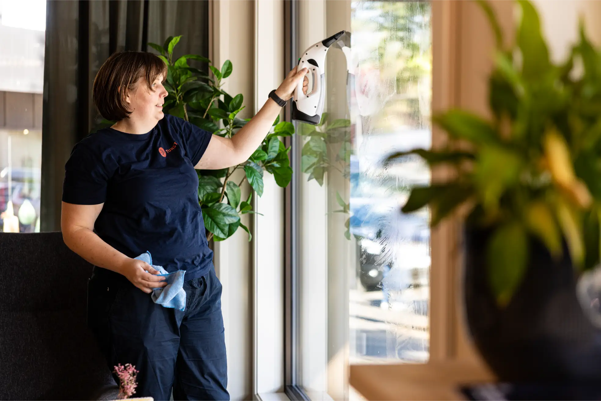 En person i mørke klær, som muligens søker en ledig stilling som arbeidsleder, vasker et vindu med et rengjøringsredskap i den ene hånden og en klut i den andre. Det står grønne planter i nærheten.